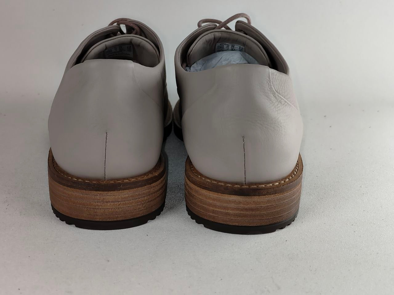 A pair of light gray leather lace-up shoes shown from the back, featuring a stacked brown wood-look heel and sole, on a light gray background.