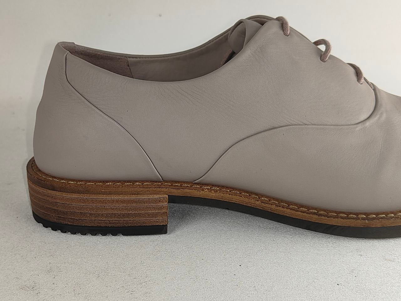 A light taupe leather lace-up oxford shoe with a stacked wood-look heel and a black outsole, shown from a side profile view against a neutral light gray background.