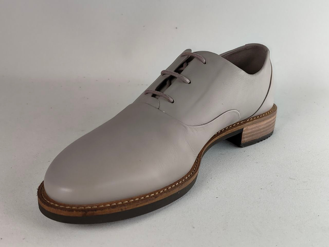 A single men's oxford dress shoe in a light taupe-gray leather, featuring a stacked wood-look heel and a dark brown sole, shown against a neutral light gray background.