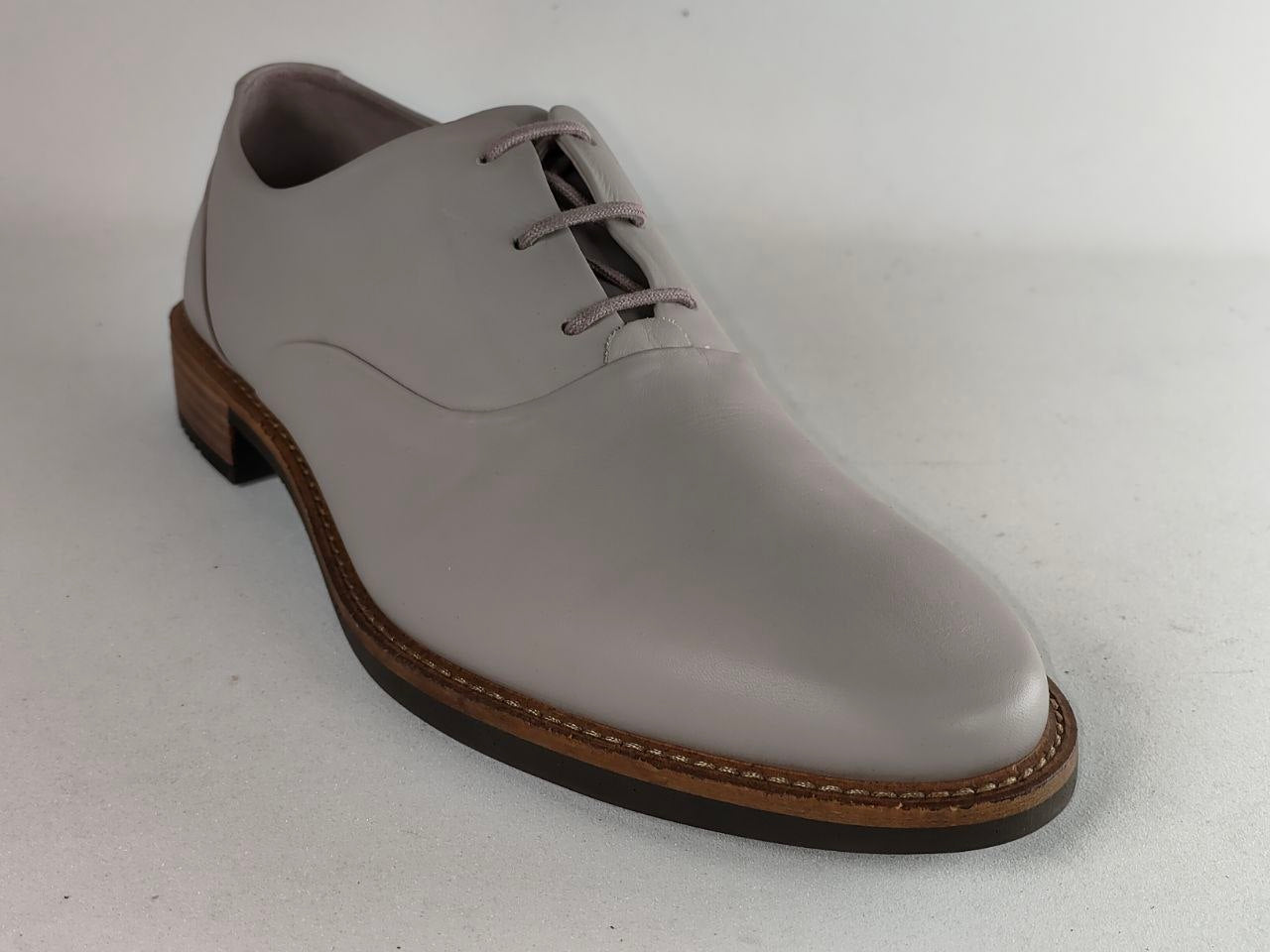 A light gray leather lace-up dress shoe with a brown stacked sole, photographed on a neutral light gray background.