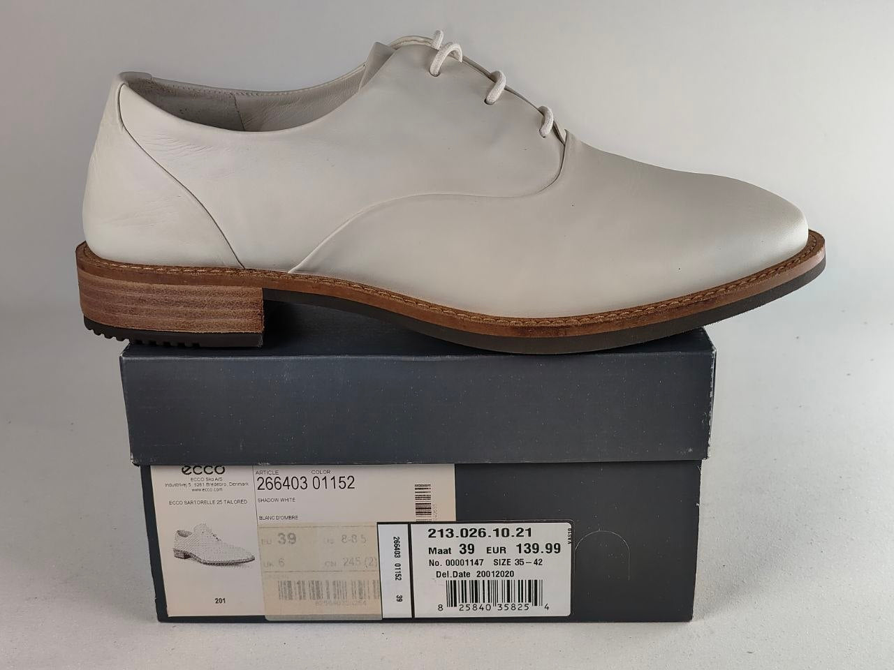 A white leather Ecco lace-up oxford shoe with a brown stacked block heel, displayed on top of its dark gray retail box.