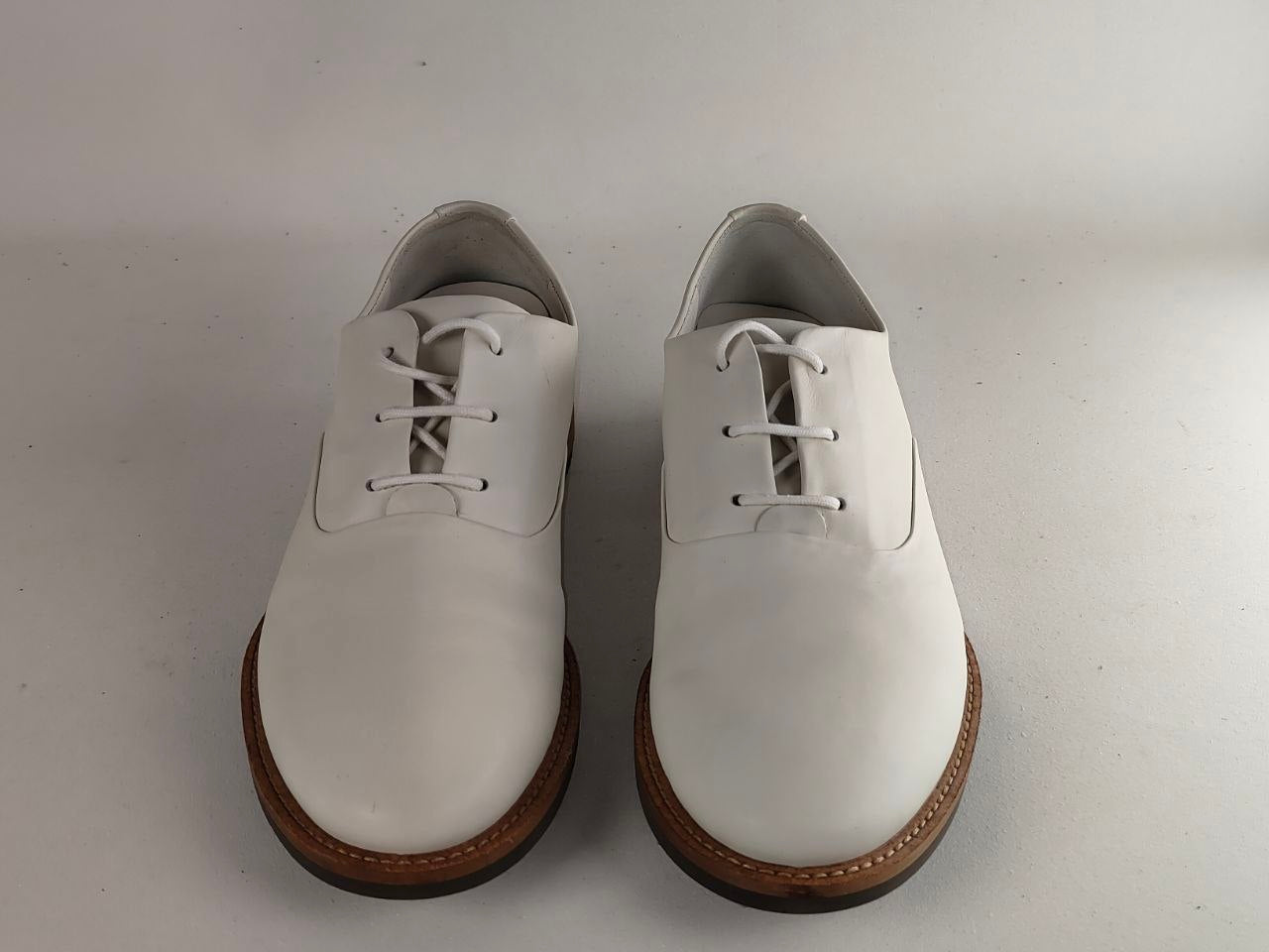A pair of white leather lace-up oxford shoes with brown soles, photographed from the front on a plain light gray background.