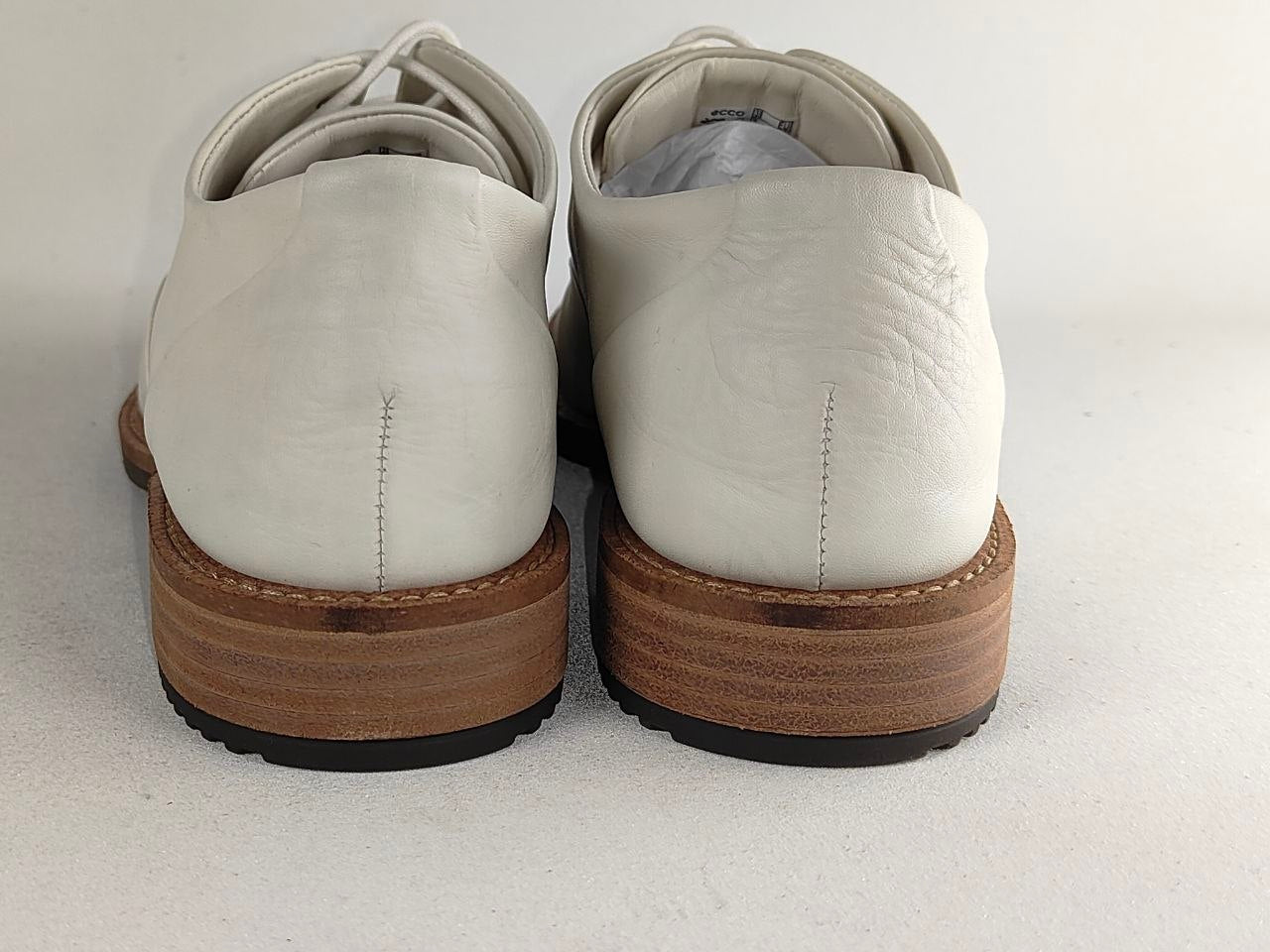 A pair of white leather lace-up shoes shown from the back, featuring a stacked brown wood-look heel and a black outsole, set against a plain light gray background.