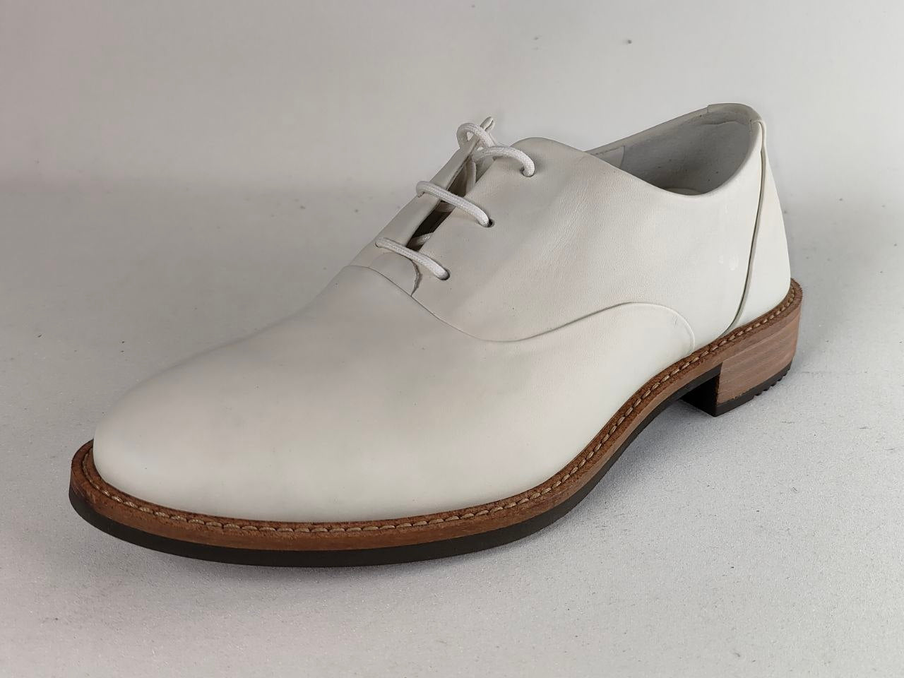 A white leather lace-up dress shoe with a brown stacked heel and welt, photographed on a light gray background.