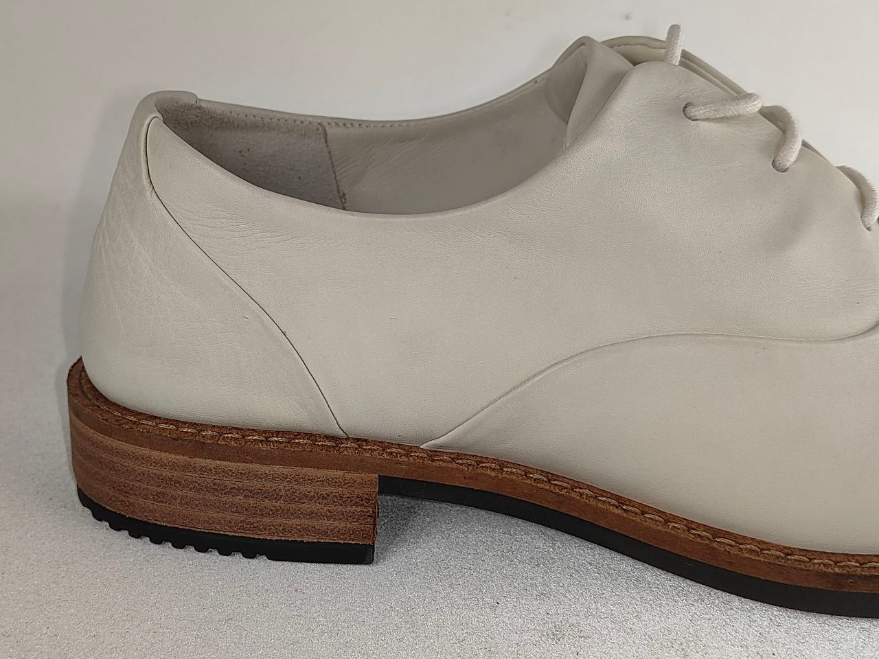 A close-up side view of a light beige leather lace-up shoe with a stacked brown wooden heel and black outsole, set against a plain light gray background.