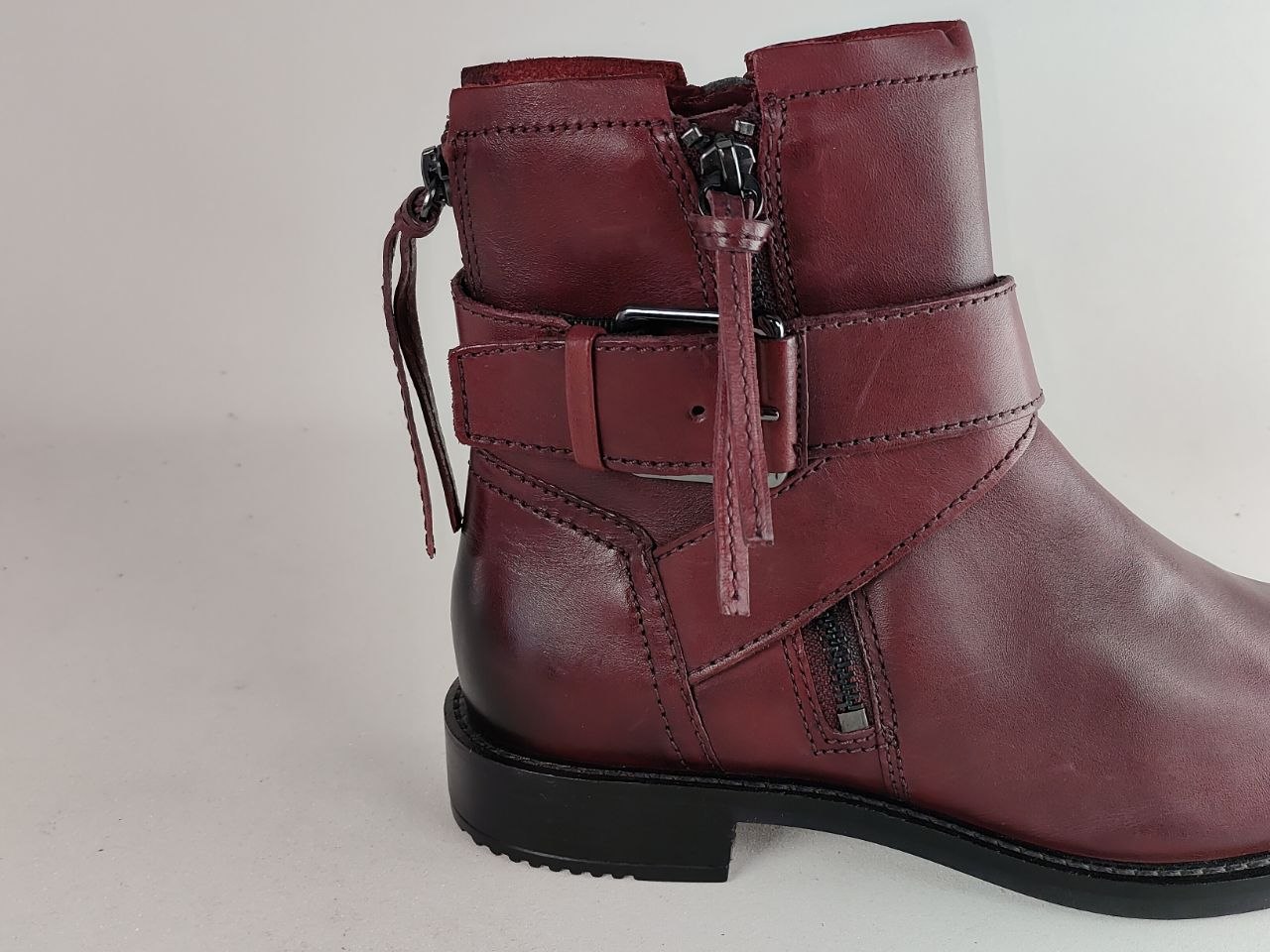 A burgundy leather ankle boot with a side zipper, a decorative strap with a buckle, and hanging leather tassels, shown against a plain light background.