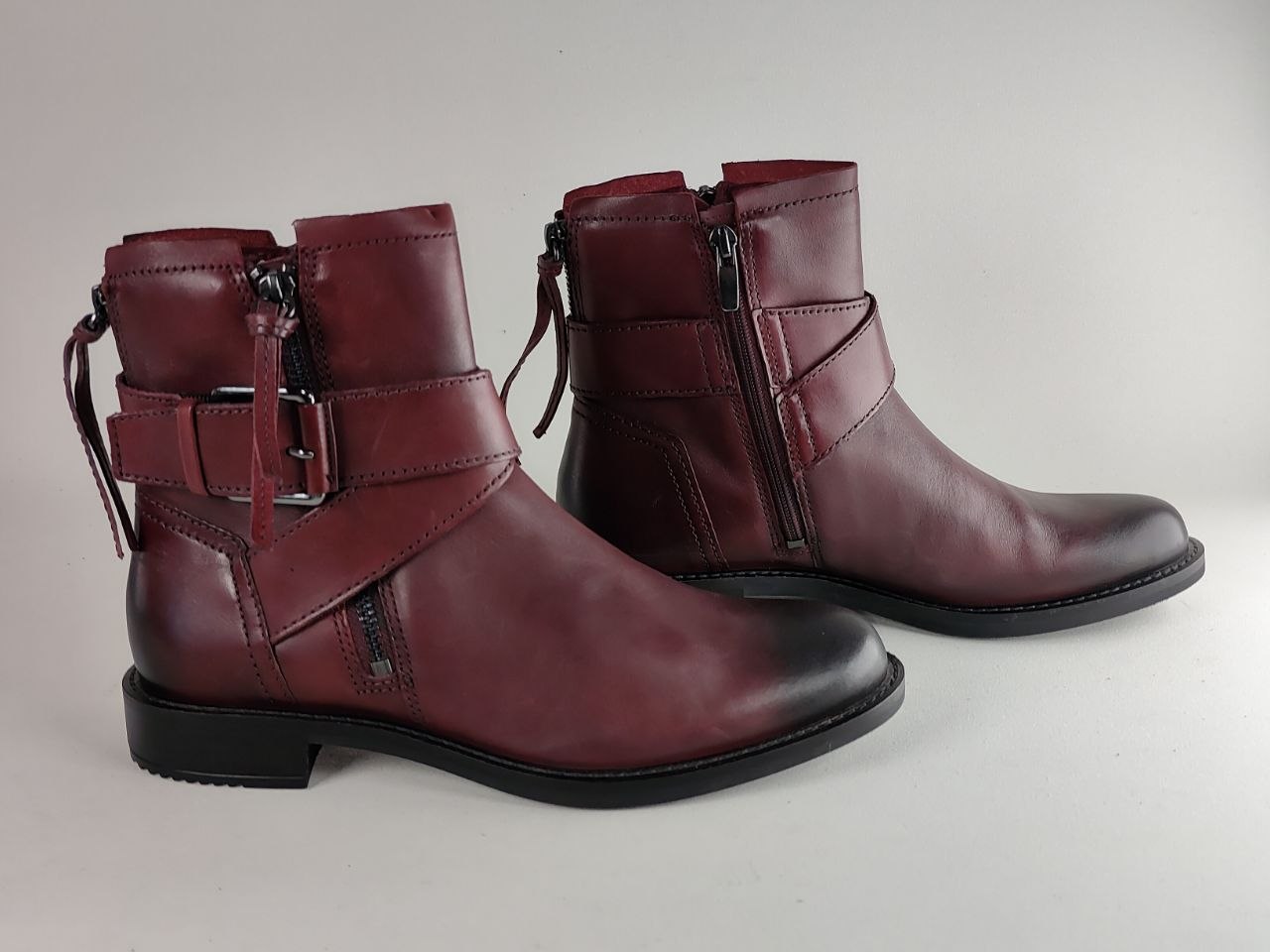 A pair of women's burgundy leather ankle boots with side zippers, buckle straps, and decorative straps on the sides, shown against a neutral light gray background.