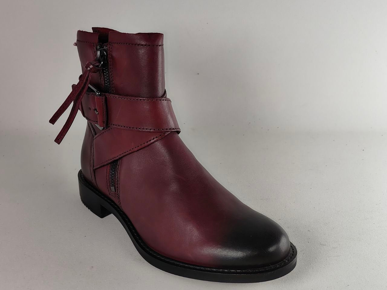 A single dark burgundy leather ankle boot with a side zipper, decorative strap with a buckle, and a matching hanging tie detail, shown against a plain light gray background.
