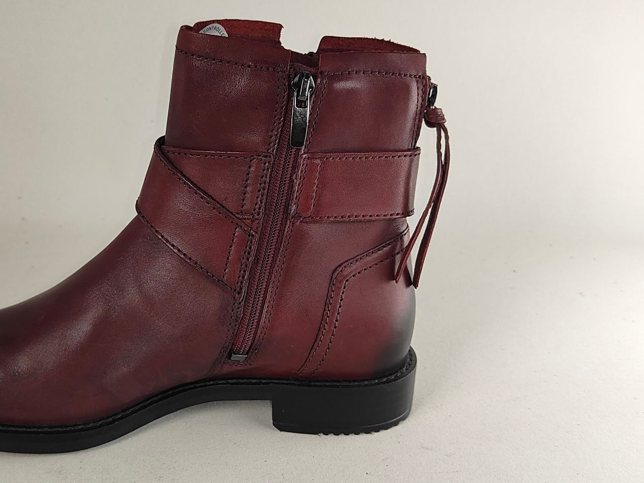A side-profile view of a dark burgundy leather ankle boot with a side zipper, decorative strap, and pull tab, set against a plain light background.