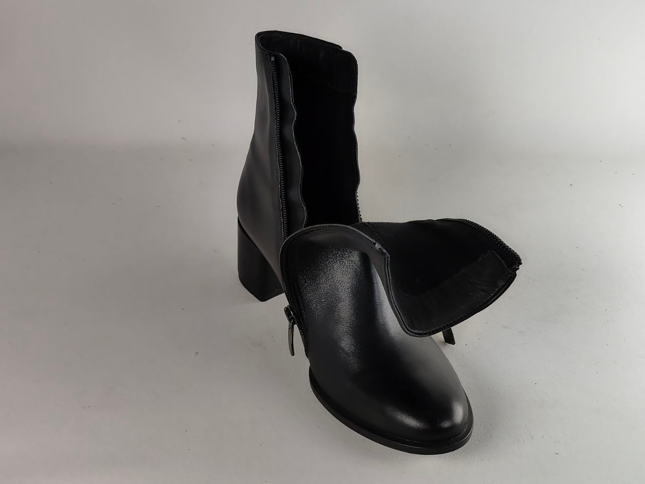 A pair of black leather ankle boots with a block heel and side zipper, photographed on a neutral light gray background.