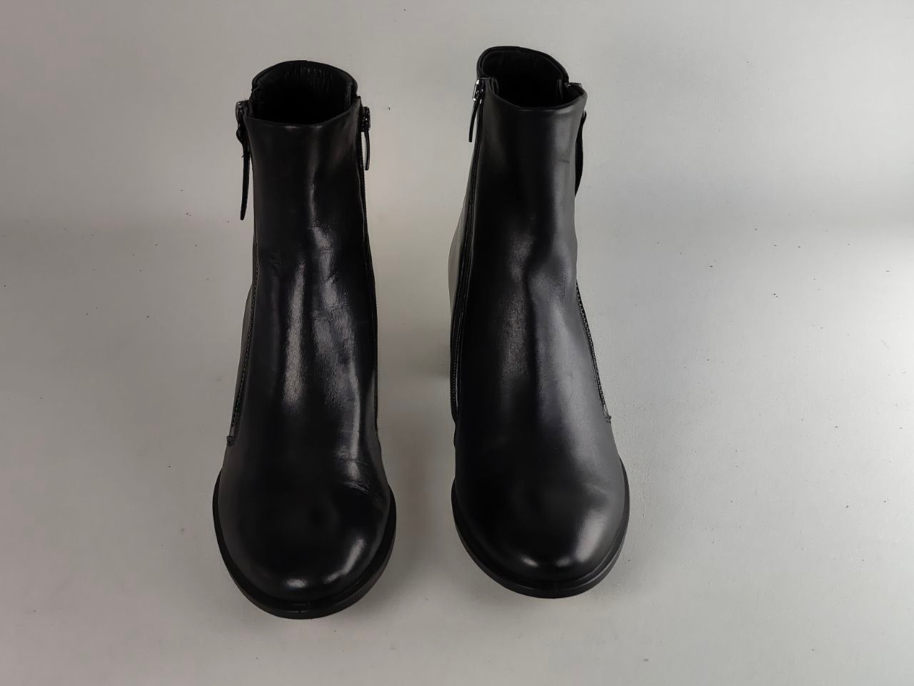 A pair of black leather ankle boots with side zippers, shown from a front-facing perspective on a neutral light gray background.