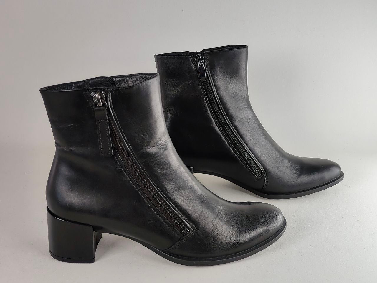 A pair of black leather ankle boots with side zippers and block heels, photographed on a neutral light gray background.