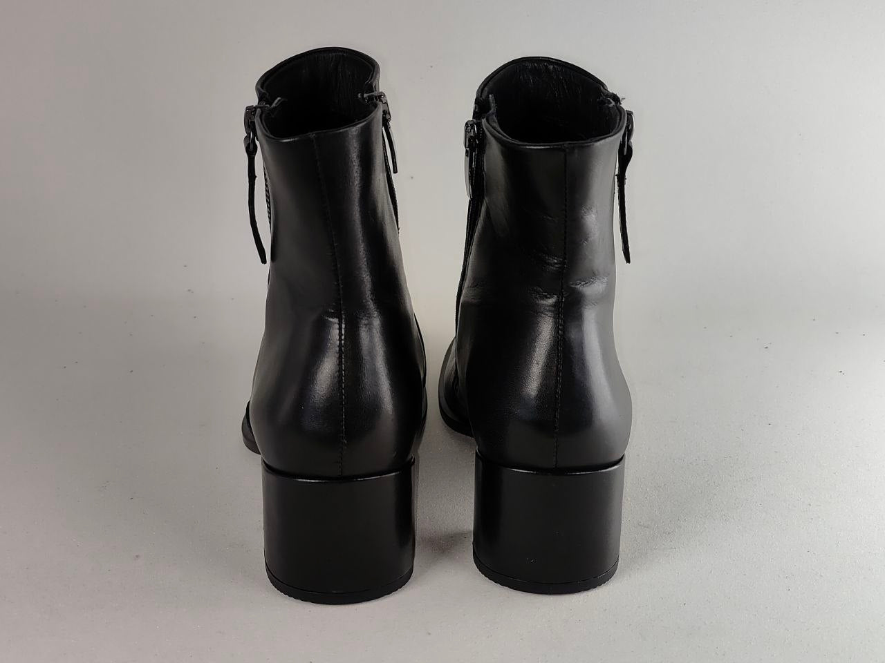 A pair of black leather ankle boots with side zippers and block heels, shown from the back against a neutral light gray background.