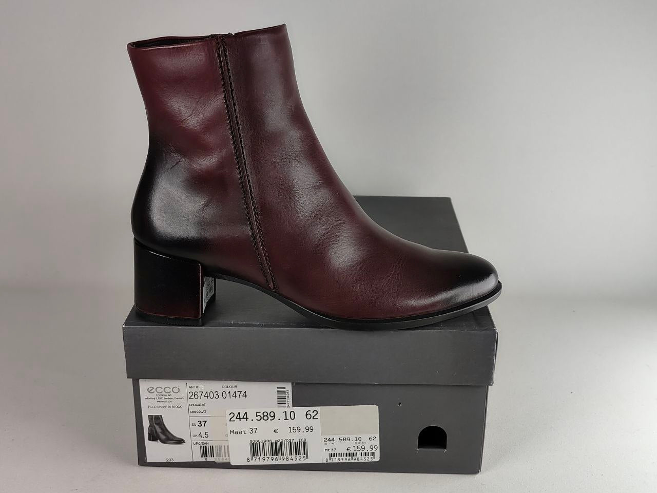 A dark burgundy leather ankle boot with a block heel, shown resting on top of its black shoebox against a neutral gray background.