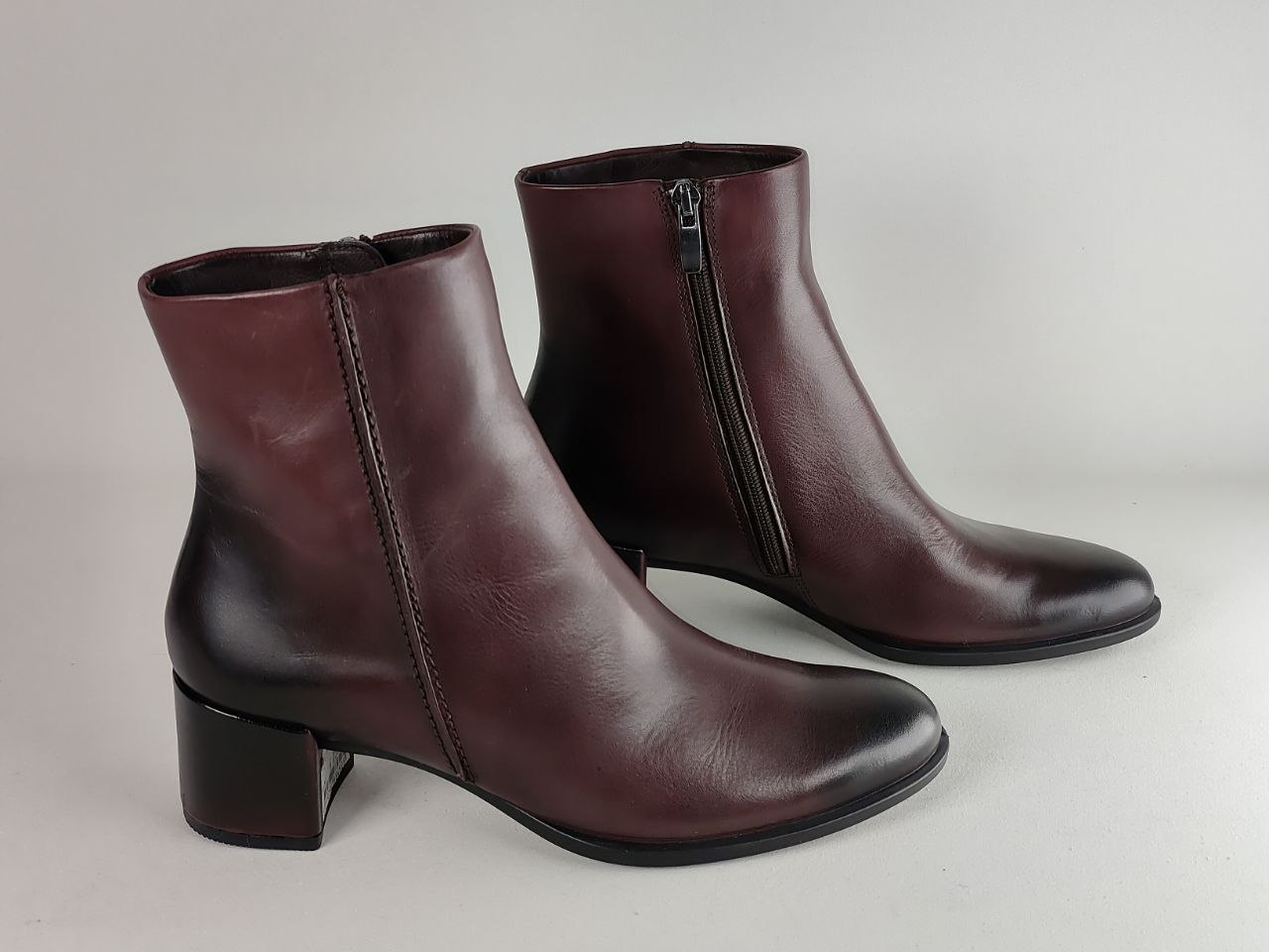 A pair of dark burgundy leather ankle boots with side zippers and low block heels, photographed on a neutral light gray background.