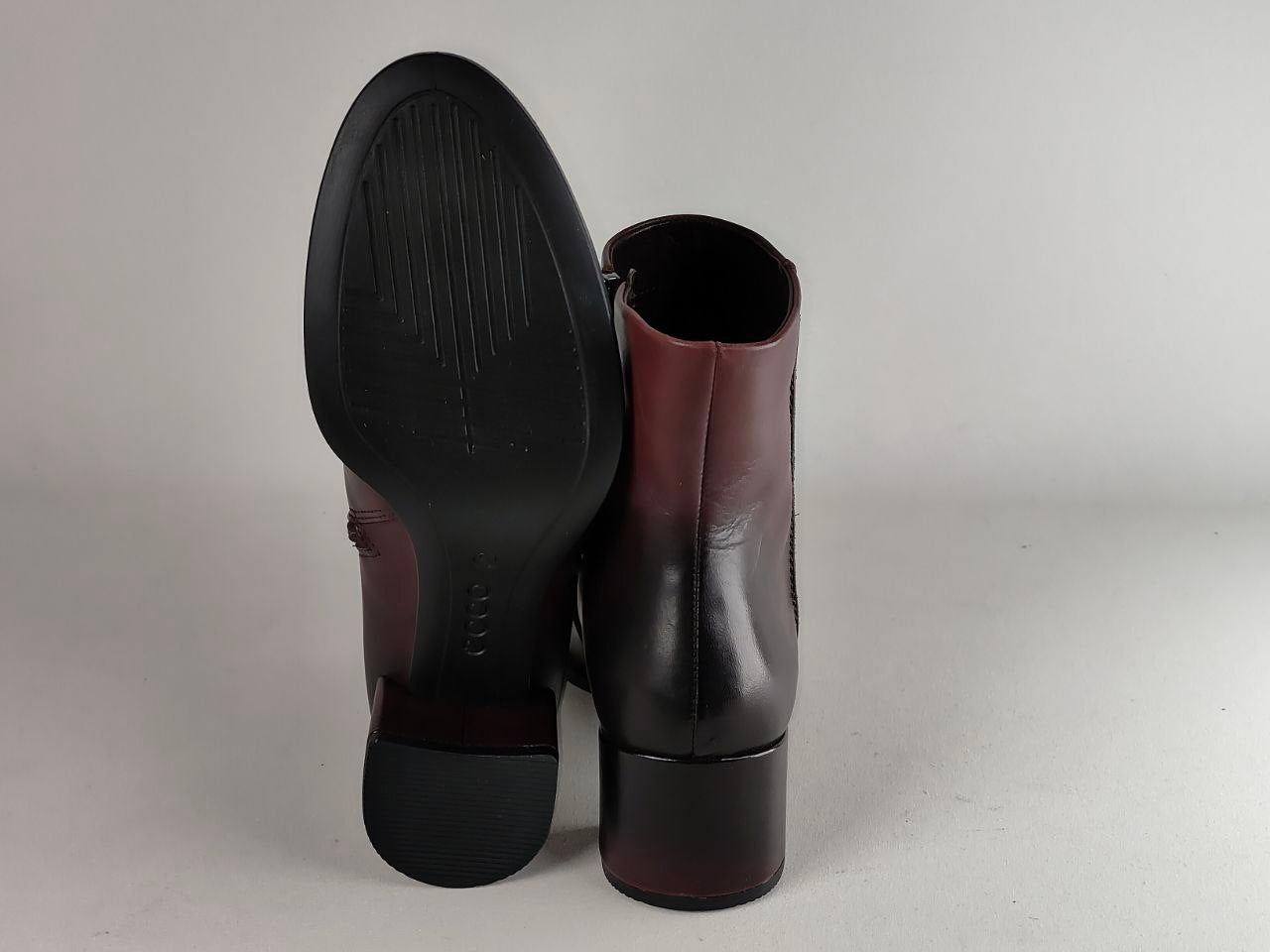 A pair of dark brown leather ankle boots shown from the back, with one boot standing upright to display the heel and sole and the other leaning against it.