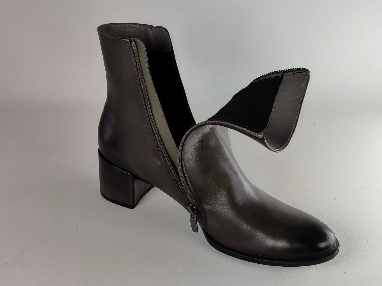 A dark brown leather ankle boot with a low block heel, featuring a side zipper and a matching pull tab, photographed on a neutral light gray background.