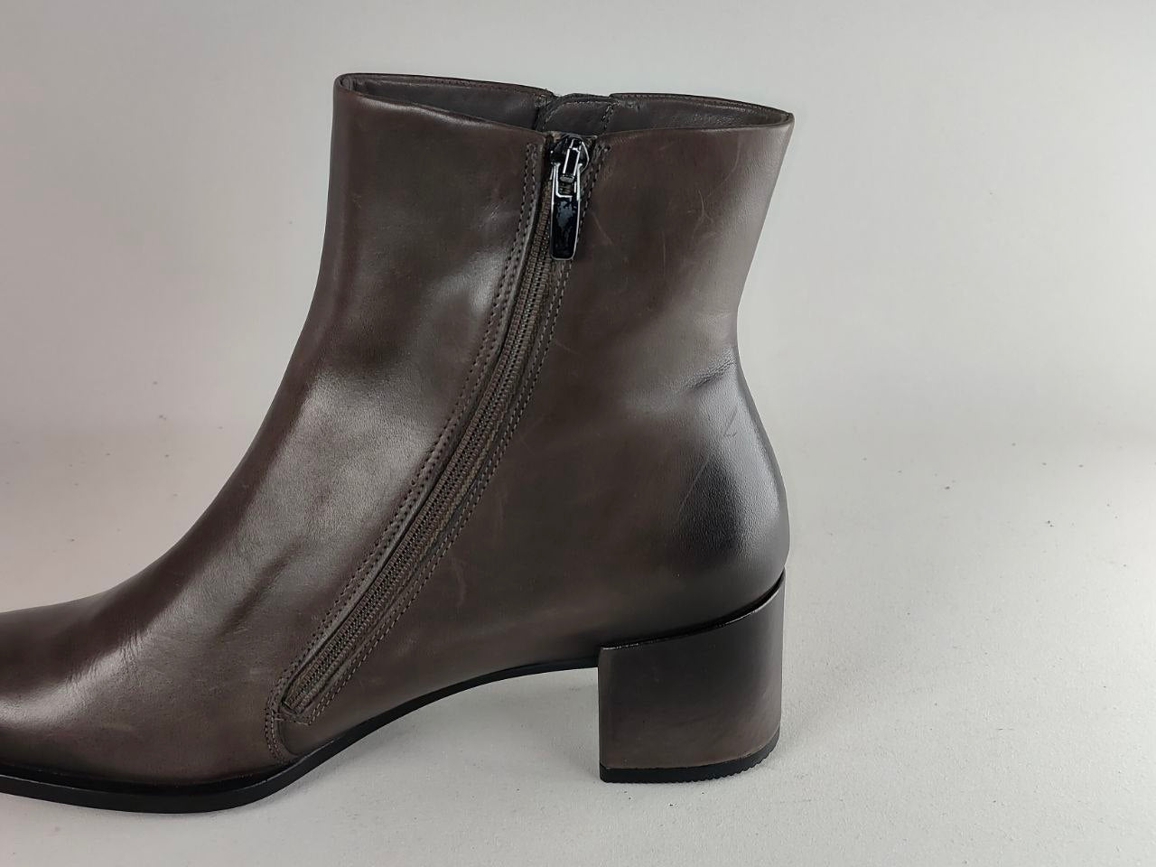 A dark brown leather ankle boot with a side zipper and a low block heel, shown from a side profile view against a neutral light gray background.