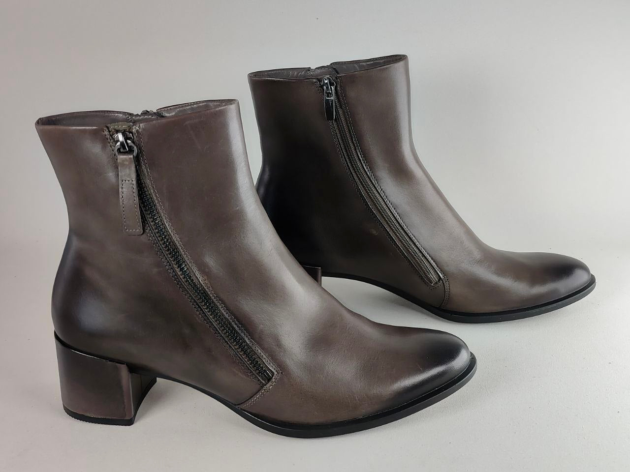 A pair of women's dark brown leather ankle boots with side zippers, low block heels, and pointed toes, shown on a neutral light gray background.