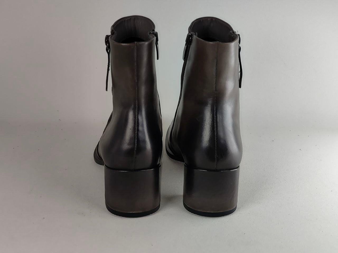 A pair of black leather ankle boots with low block heels, shown from the back against a neutral gray background.