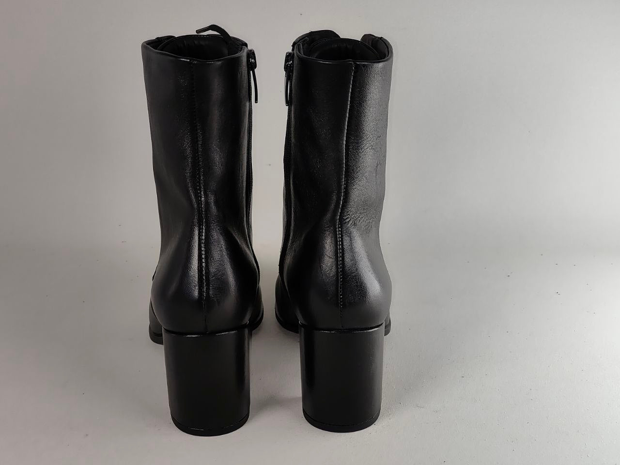 A pair of black leather ankle boots with block heels, shown from the back against a neutral gray background.