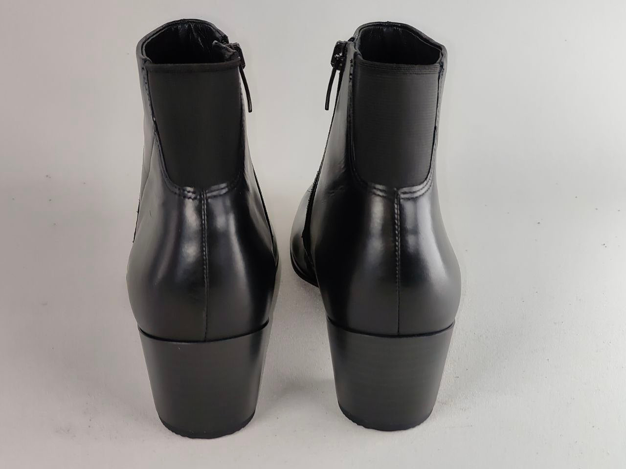 A pair of black leather ankle boots with side zippers and block heels, shown from the back against a light gray background.