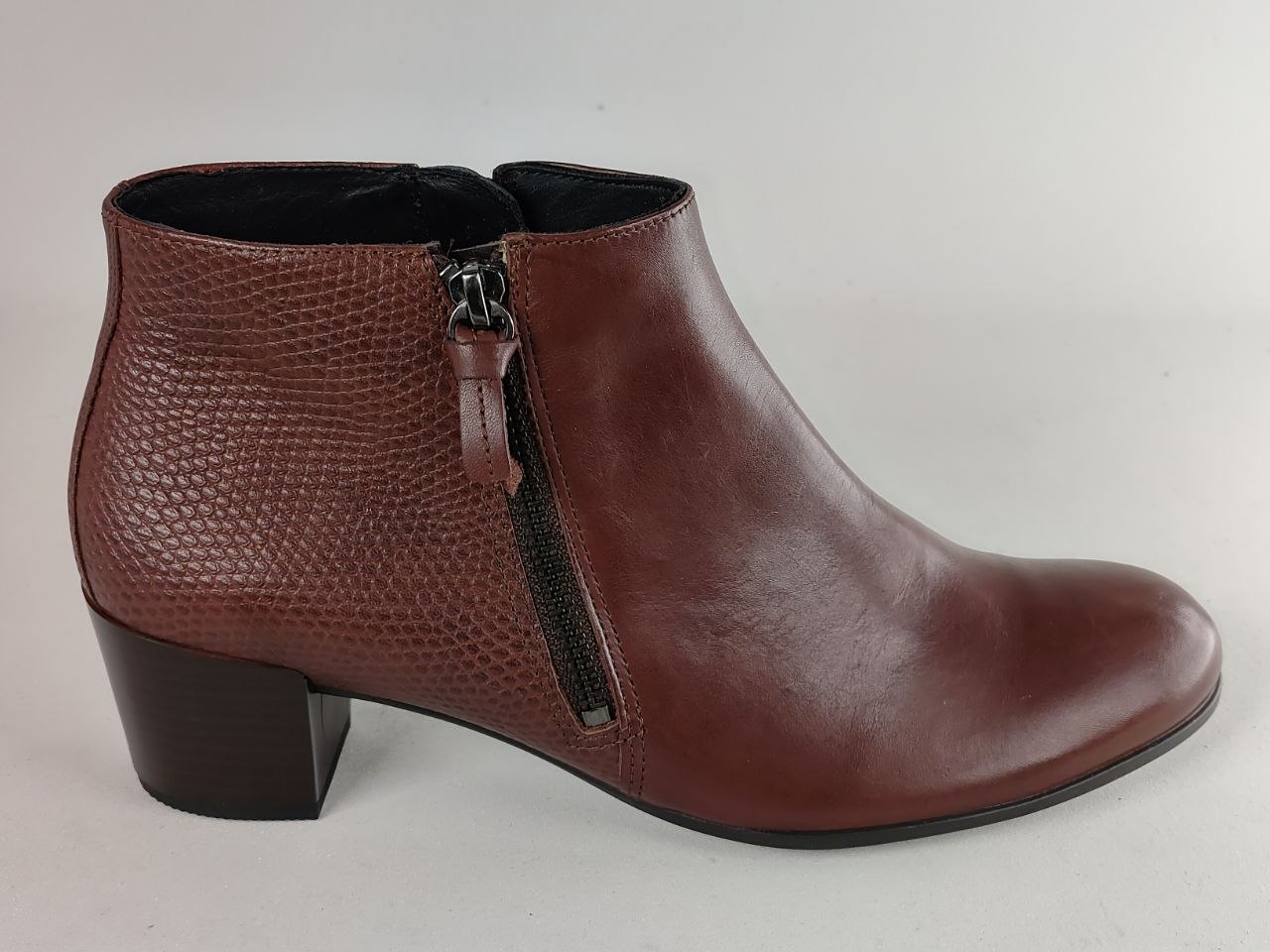 A single women's ankle boot in a reddish-brown color, featuring a textured crocodile-patterned side panel, smooth leather on the toe and heel, and a black side zipper with a pull tab, shown against a plain light gray background.