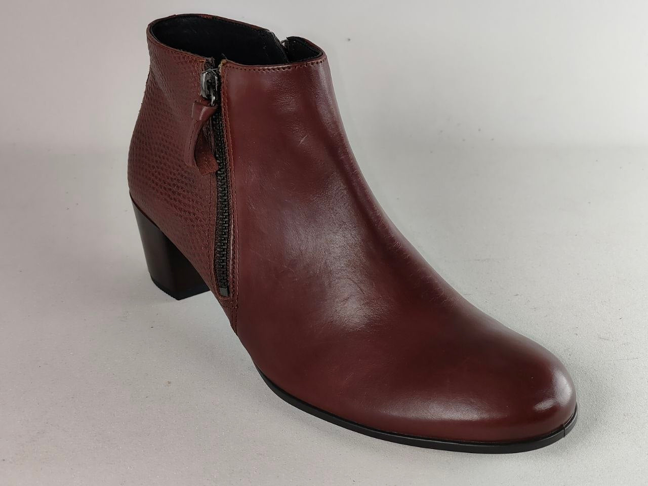 A single women's ankle boot in a deep burgundy-brown leather, featuring a side zipper, a small decorative pull tab, and a low block heel, shown against a neutral light gray background.