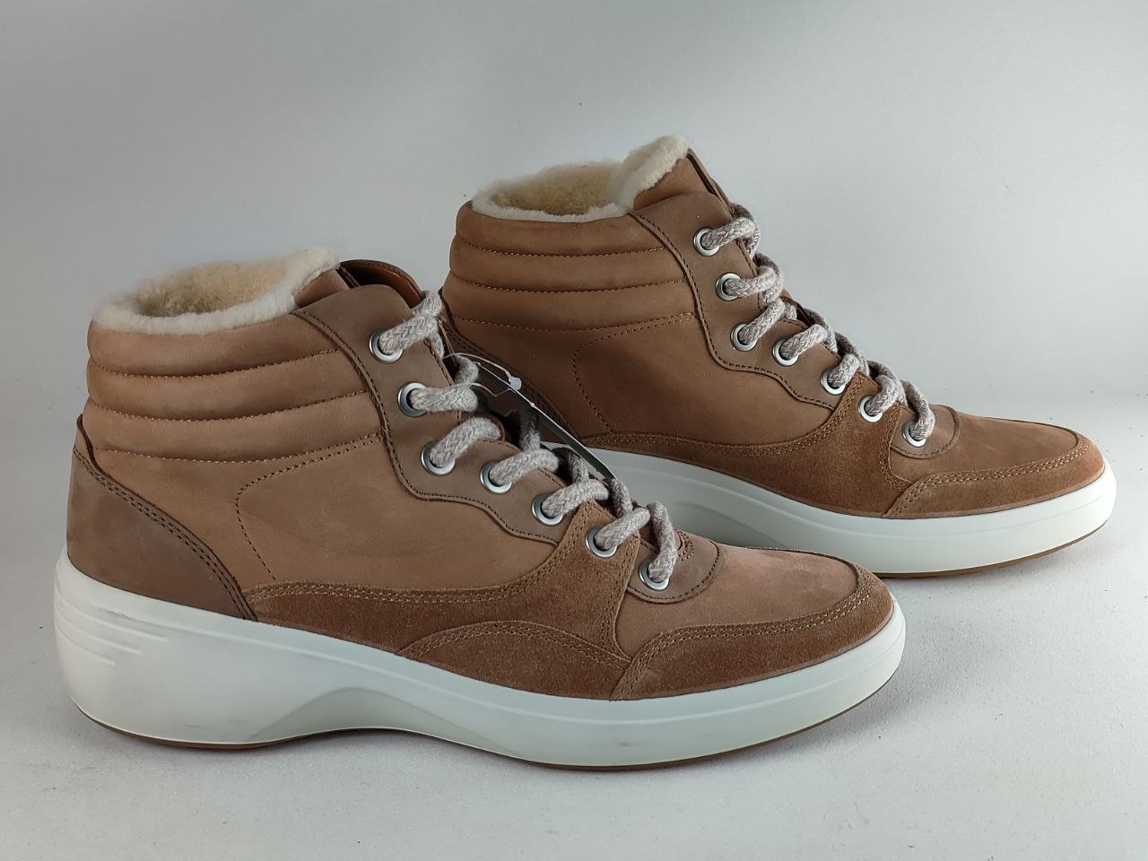 A pair of mid-height brown suede sneakers with white shearling-style lining, white laces, and thick white platform soles, photographed on a light gray studio background.