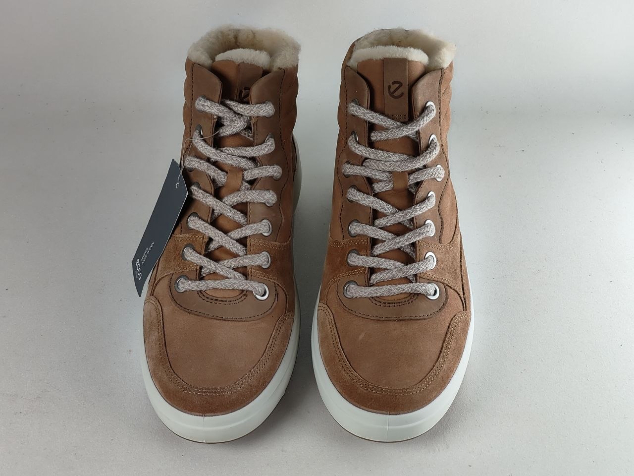 A pair of brown suede high-top sneakers with white laces, white soles, and a soft beige shearling lining, shown against a light gray background.