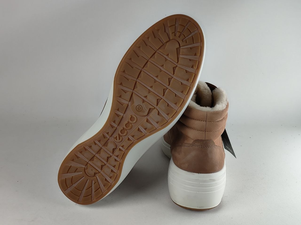 A pair of mid-calf boots shown on a light gray studio background, with one boot tilted to display the brown outsole and the other standing upright to show the tan leather upper, white sole, and white faux-fur collar.