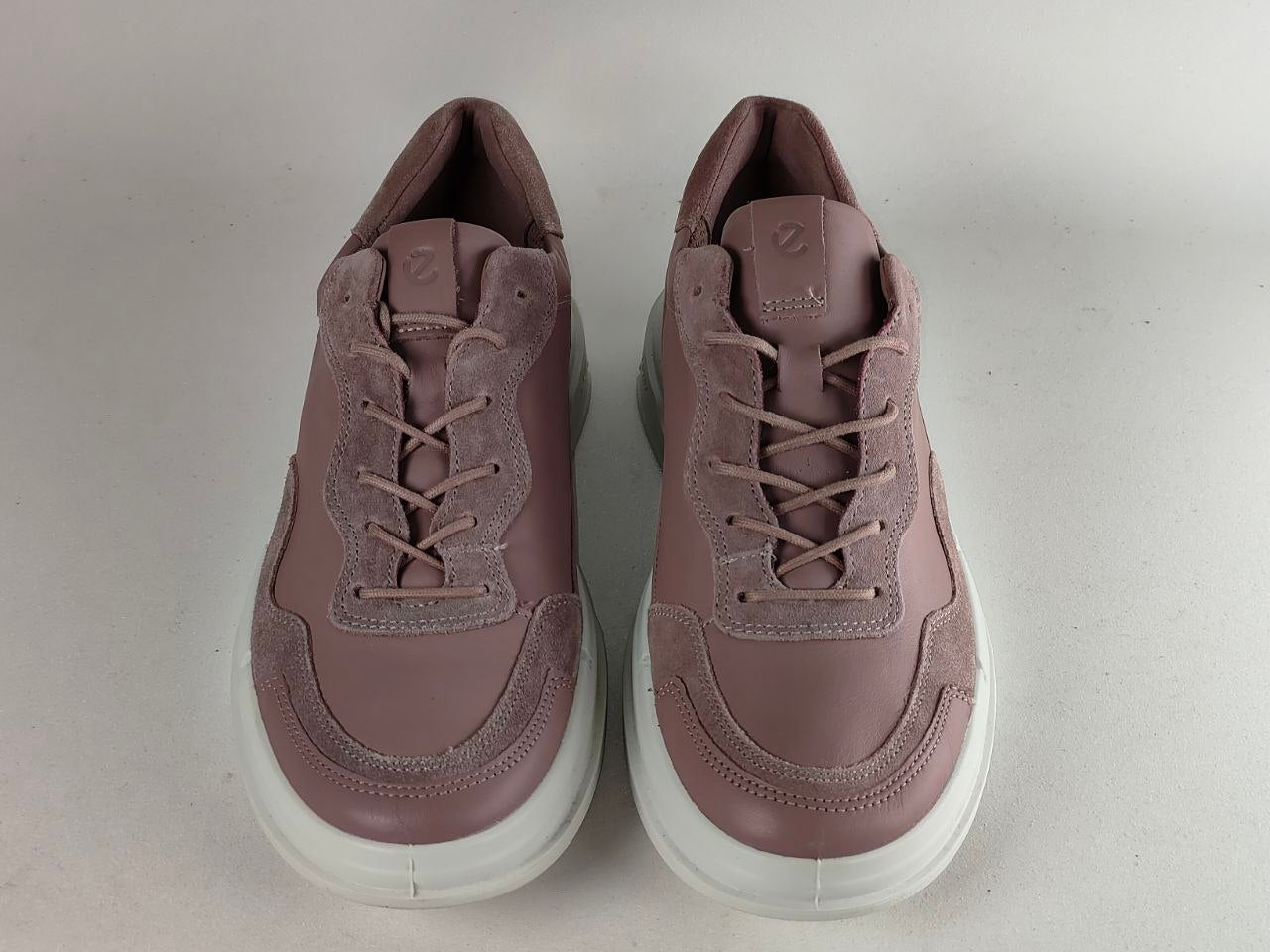 A pair of low-top lace-up sneakers in a dusty mauve or muted rose color, featuring a mix of smooth and suede-like textures, set against a plain light gray background.