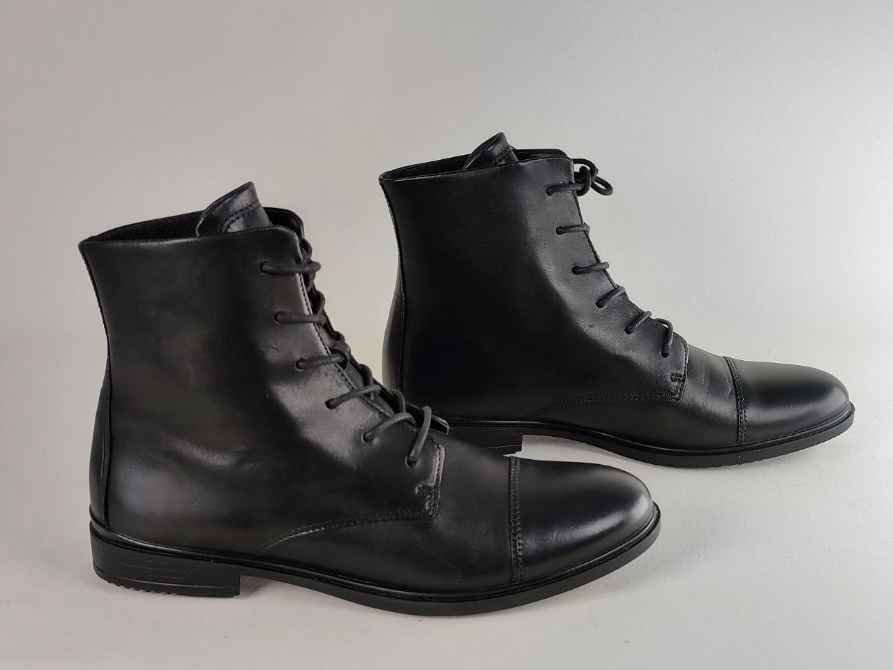 A pair of black leather lace-up ankle boots with a cap toe, photographed on a neutral light gray background.