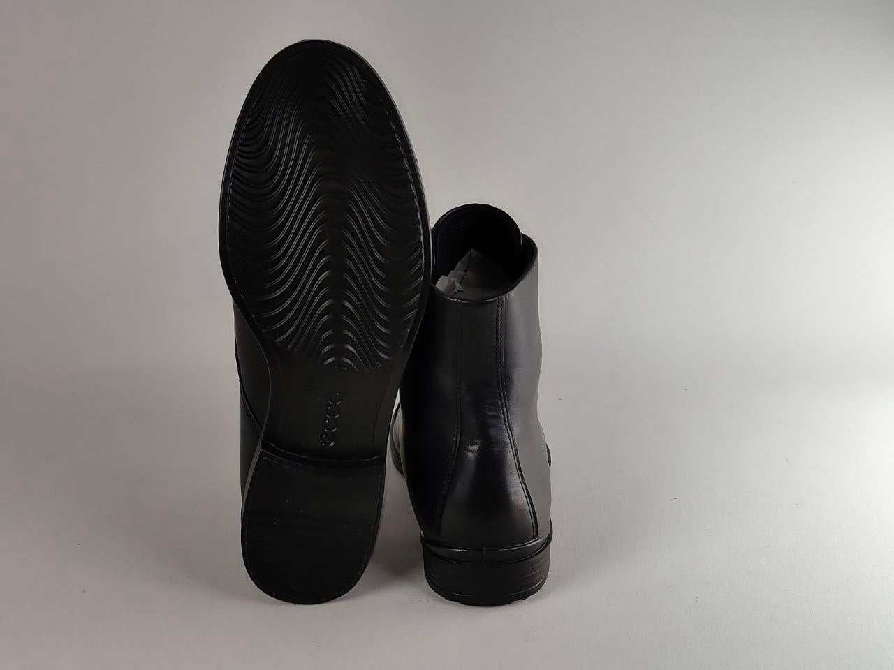 A pair of black leather ankle boots shown from the back, with one boot standing upright to display the textured black rubber sole and the other lying flat to show the back profile.