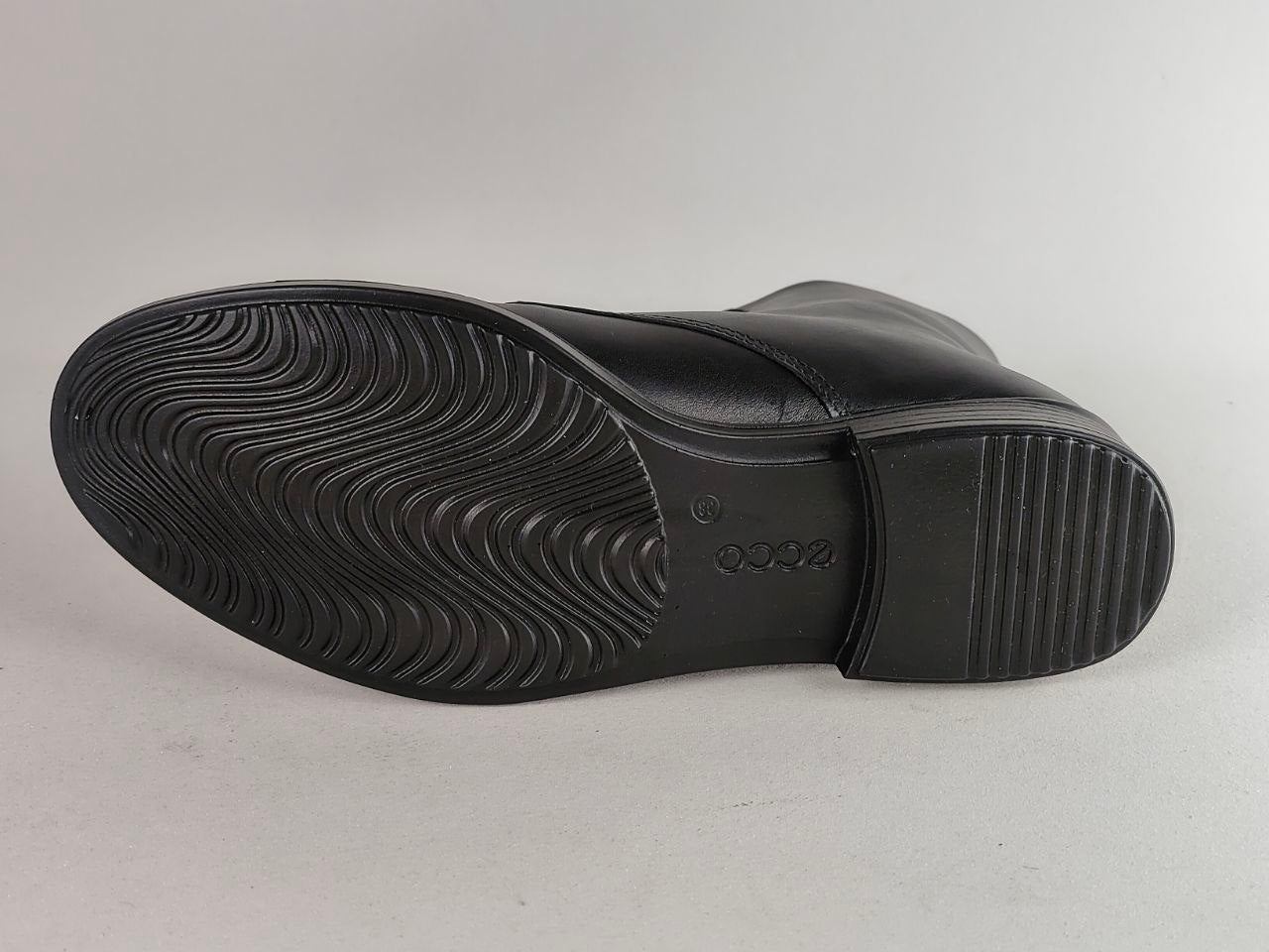 The black rubber outsole of a leather shoe, showing a wavy tread pattern and embossed branding, photographed on a neutral light gray background.