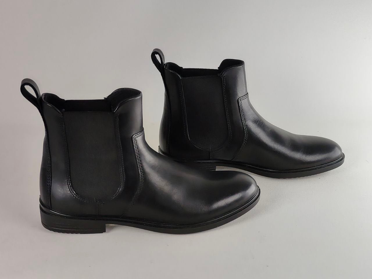 A pair of black leather Chelsea boots with elastic side panels and pull loops, photographed on a neutral light gray background.