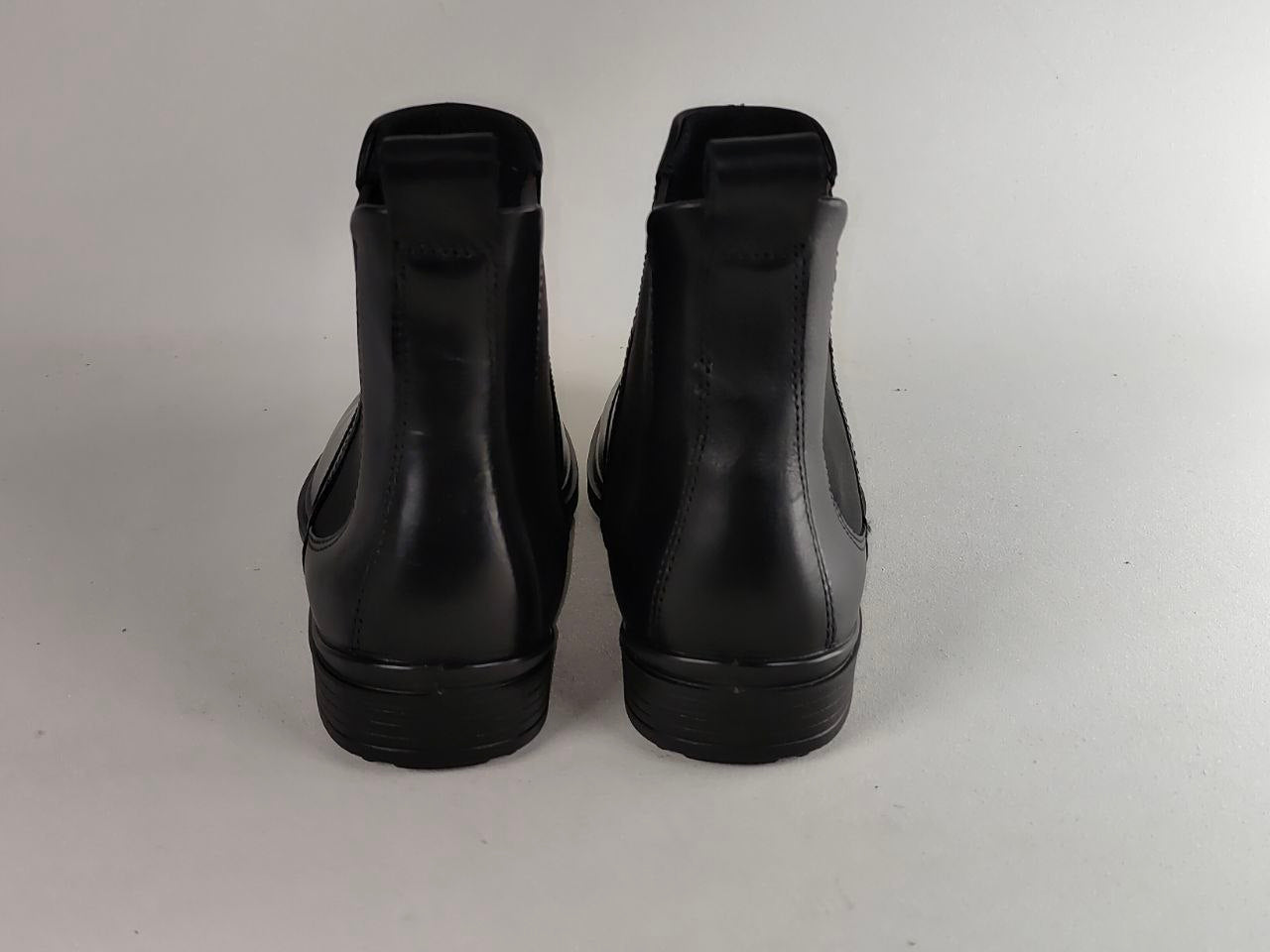 A pair of black leather Chelsea boots shown from the back, positioned side-by-side against a neutral light gray background.