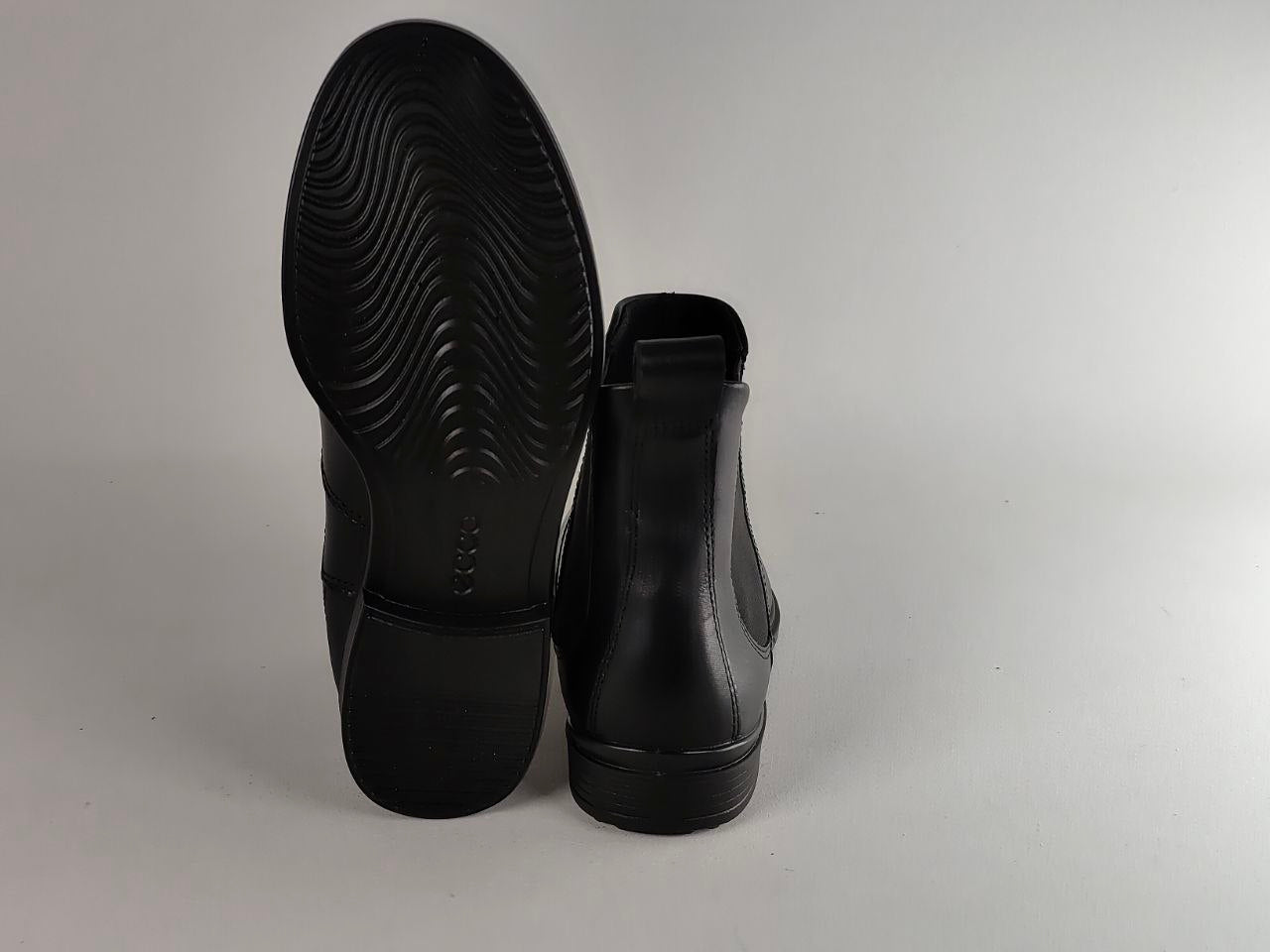 A pair of black leather Chelsea boots shown from the back, with one boot standing upright to display the textured black rubber sole and the other leaning against it, set against a plain light gray background.