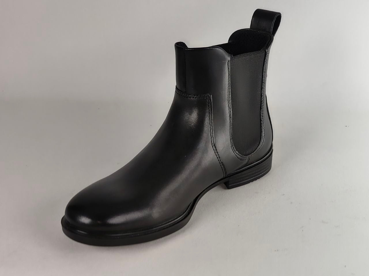 A single black leather Chelsea boot with elastic side gore panels and a pull tab, photographed on a neutral light gray background.