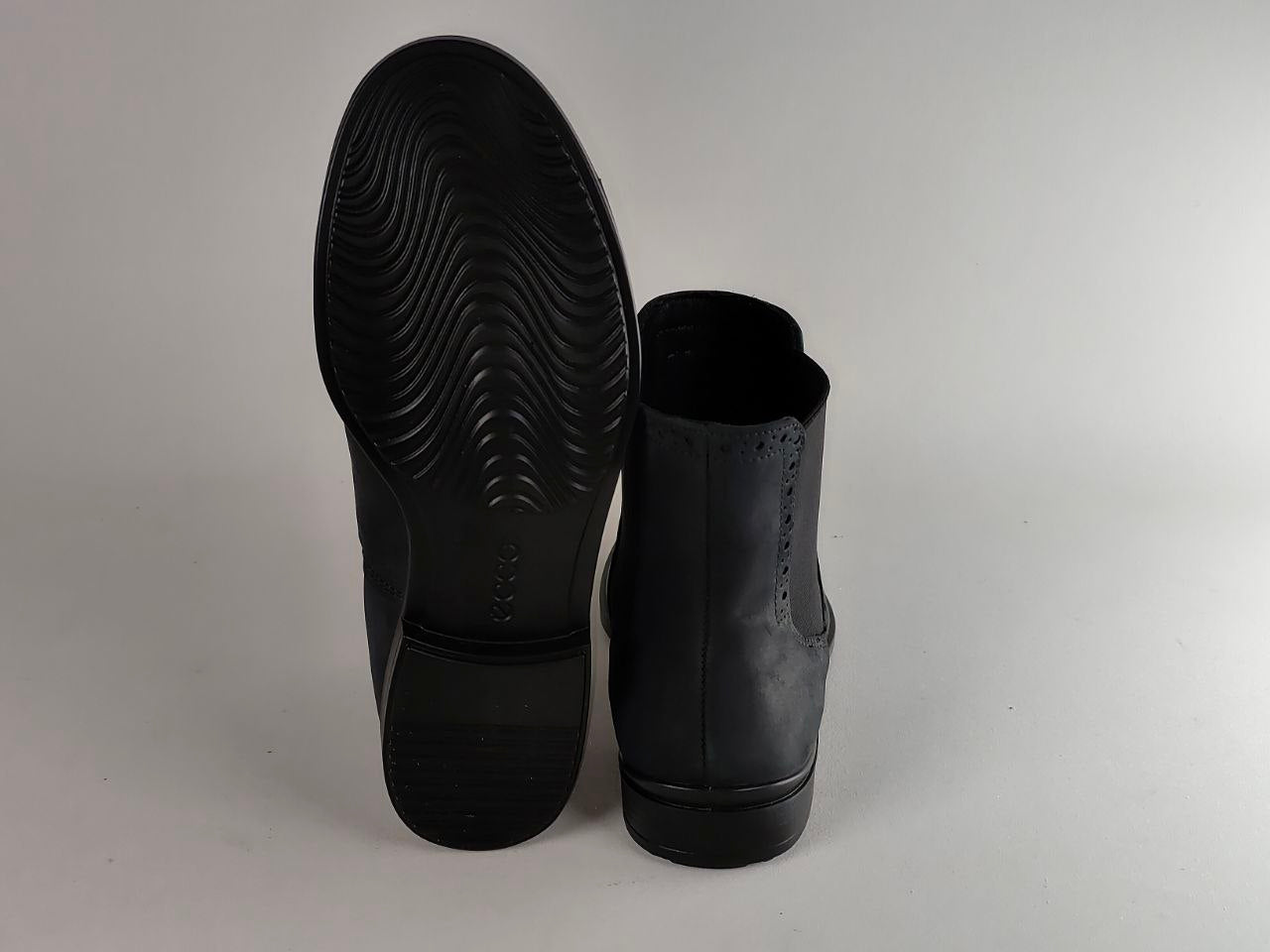 A pair of black leather ankle boots shown from the back, with one boot standing upright to display the heel and sole tread and the other lying flat to show the back profile.