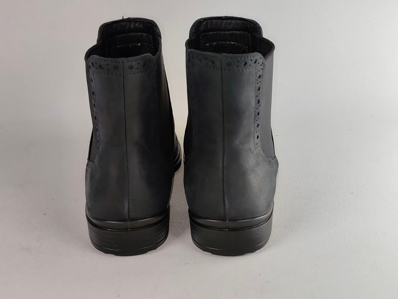 A pair of black leather ankle boots shown from the back, featuring decorative perforated detailing near the collar and thick black soles, set against a plain light gray background.