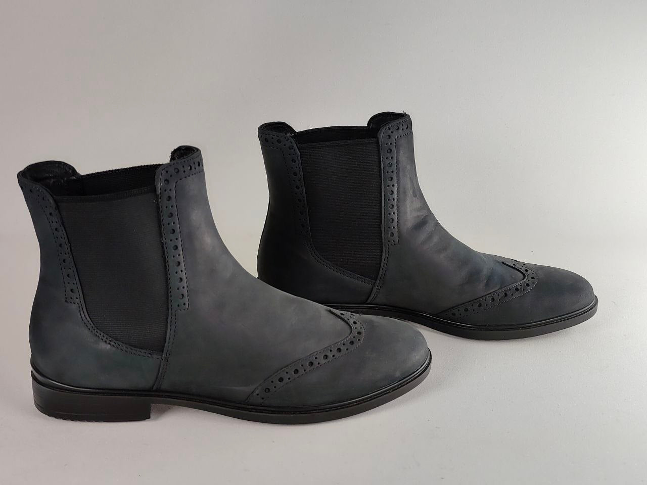 A pair of black leather Chelsea-style ankle boots with wingtip brogue detailing, shown on a neutral light gray background.