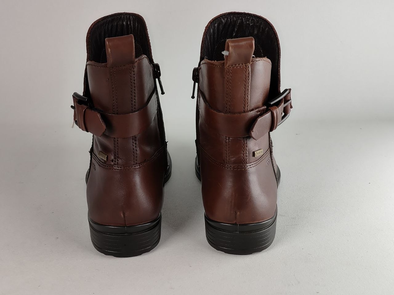 A pair of dark brown leather ankle boots with buckle straps on the sides, shown from the back against a neutral light gray background.