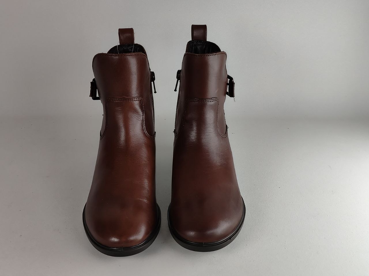 A pair of dark brown leather ankle boots with side zipper and strap details, shown from the front against a neutral light gray background.