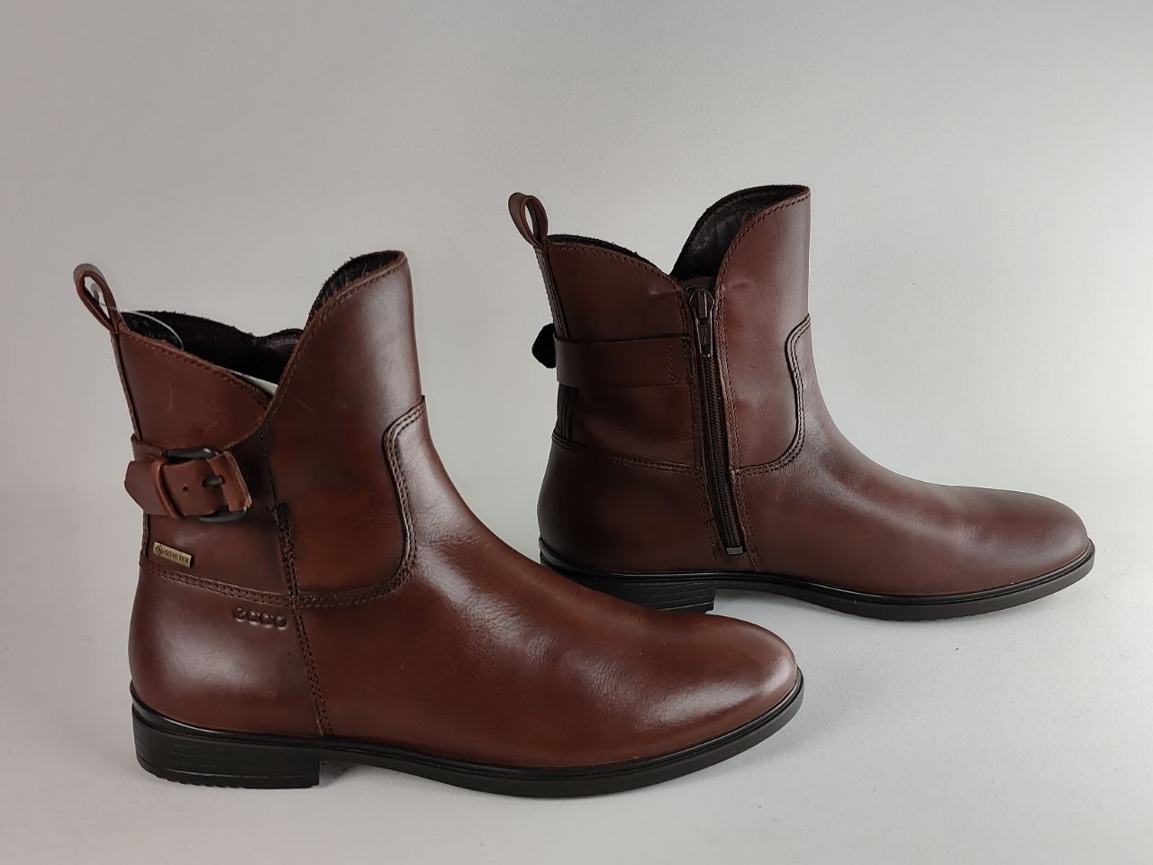 A pair of brown leather ankle boots with side zippers and buckle straps, displayed on a neutral light gray background.