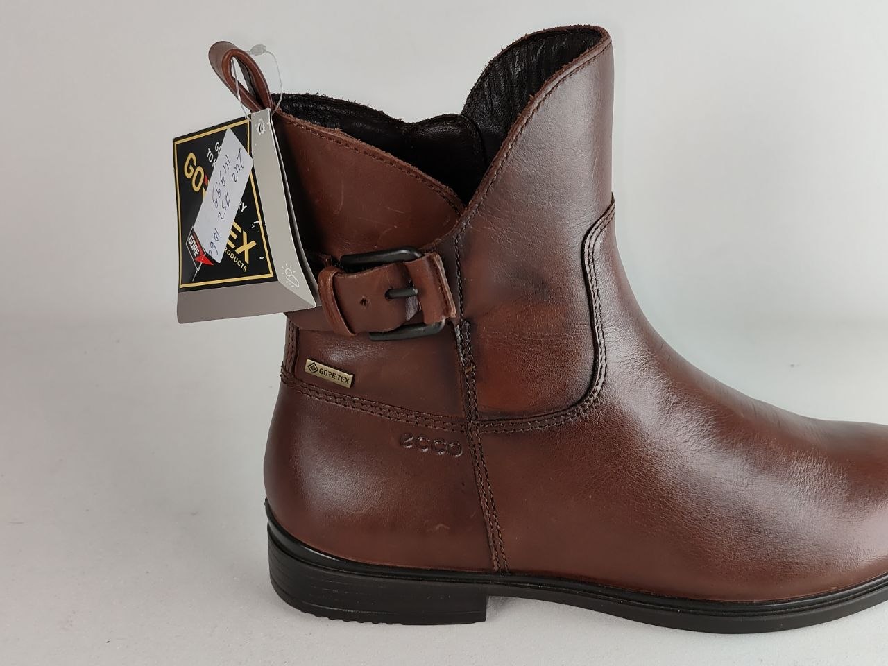 A brown leather ankle boot with a side buckle strap, a black sole, and a hangtag attached near the collar, photographed against a plain light background.
