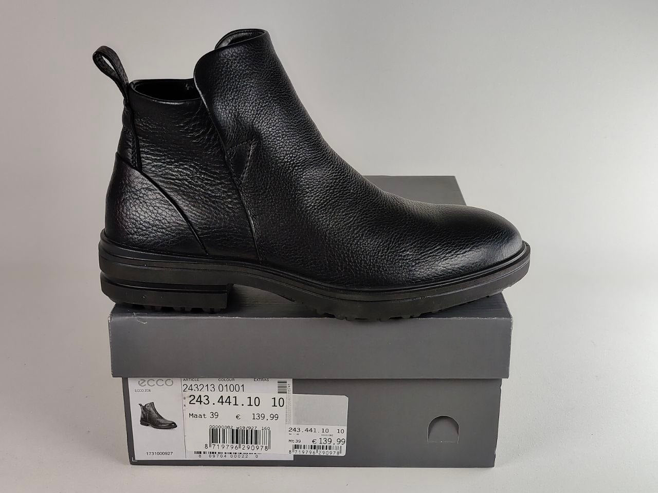 A black pebbled leather Chelsea-style ankle boot shown in profile, resting on top of a gray shoebox with a size label visible.