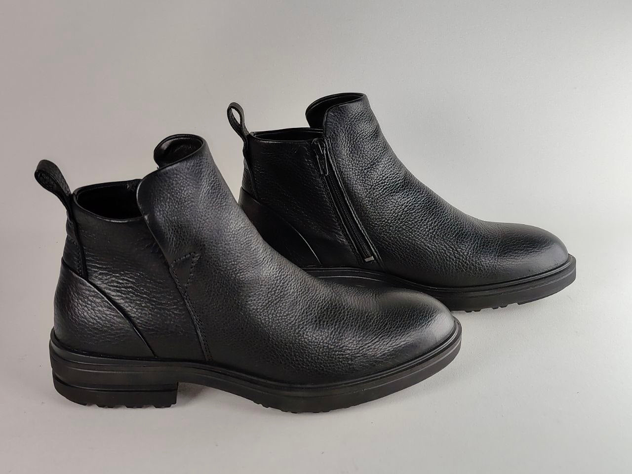 A pair of black pebbled leather ankle boots with side zippers and pull tabs, displayed on a neutral light gray background.