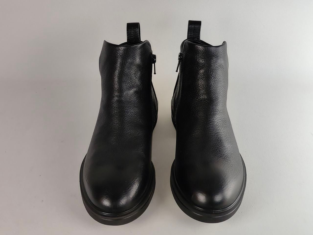 A pair of black pebbled leather ankle boots with side zippers and pull tabs, shown from the front on a light gray background.