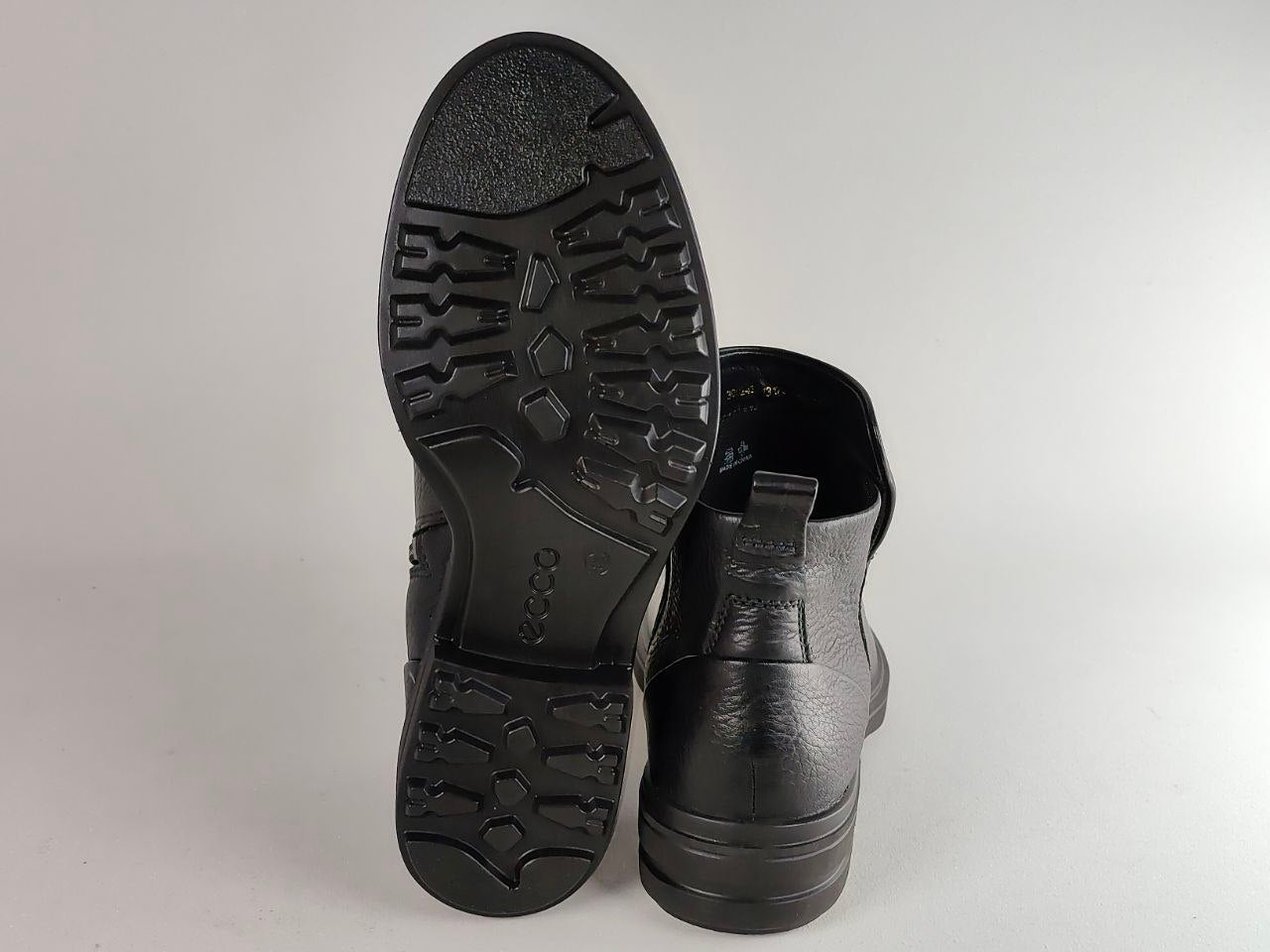 A pair of black leather ankle boots, with one boot showing the textured treaded outsole and the other standing upright to show the side profile, on a light gray background.